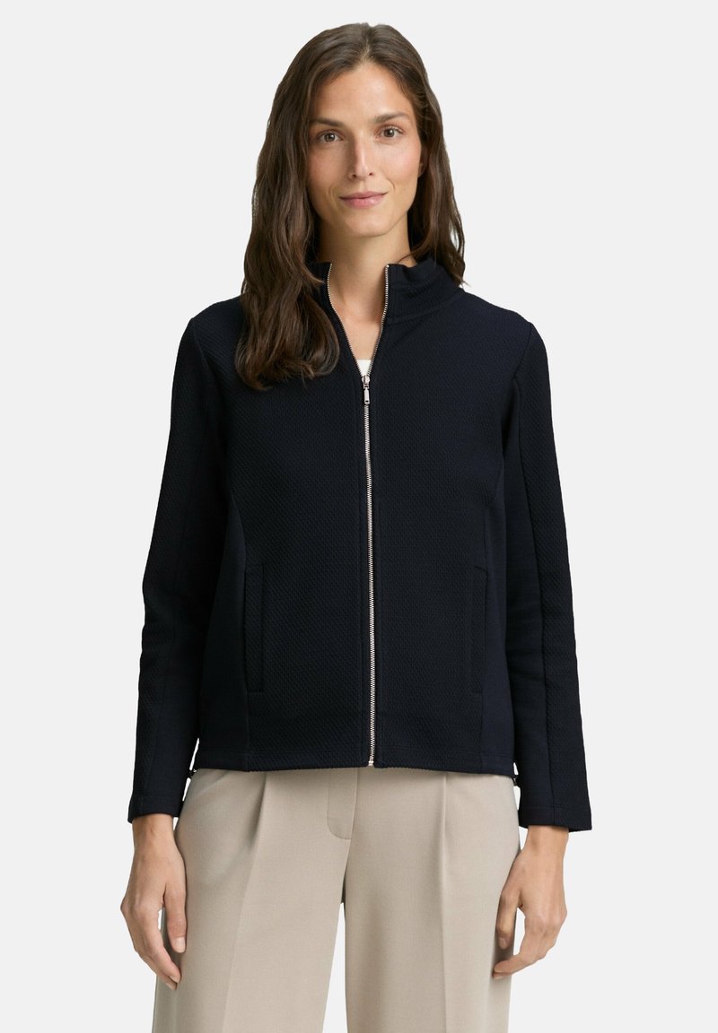Woman wearing a black zip-up textured jacket and beige pleated trousers, standing against a plain white background.
