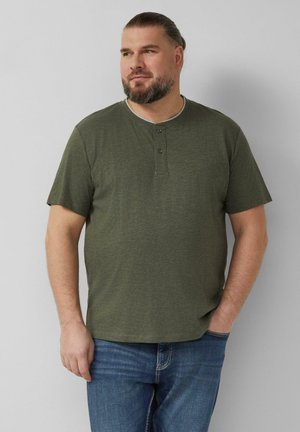 Mann mit Bart und Pferdeschwanz, der ein grünes Henley-Shirt mit kurzen Ärmeln und blaue Jeans trägt und mit einer Hand in der Tasche steht.