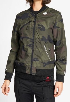 Woman wearing a green and black camo zip-up bomber jacket with black ribbed cuffs and hem, paired with black pants featuring a red logo.