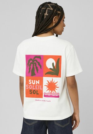 Femme aux cheveux tressés portant un t-shirt blanc avec des motifs colorés de soleil et de plante, et les mots « Sun Soleil Sol » au dos.