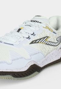 Λευκό αθλητικό παπούτσι με υφή στην επάνω επιφάνεια, μαύρο και χρυσό λογότυπο στο πλάι, λευκά κορδόνια και πουά σόλα με σκούρα καφέ πέλμα.