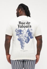 Balta kokvilnas T-kreklu ar zilu grafiku, kas attēlo vīnogas un lapas uz muguras, ar tekstu "Rue de Valouis" un "Creative Studio."