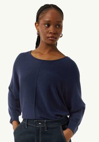 Marineblauer Strickpullover mit weitem Rundhalsausschnitt, überschnittenen Schultern und gerippten Ärmelabschlüssen. Kombiniert mit dunklen Denim-Jeans mit sichtbaren Nähten.
