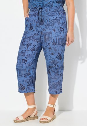 Pantalons bleus courts avec une taille élastique et cordon de serrage, présentant un motif graphique de monuments et de nourriture. Tissu doux avec une coupe décontractée. Portés avec des sandales beiges.
