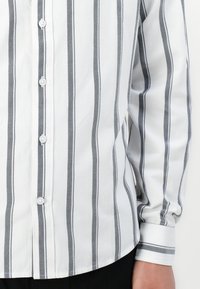 Camisa blanca de manga larga con rayas grises verticales, con botones en la parte delantera y en los puños, llevada con pantalones negros.
