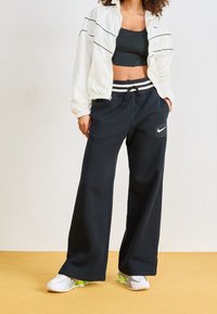 Bílé zipové sako s černými akcenty, černý crop top a volné černé tepláky s logem. Obuv má bílý základ a neonové detaily.