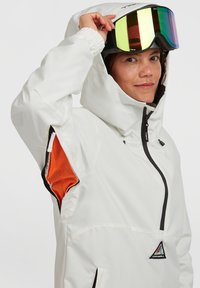 Witte waterdichte jas met een capuchon, voorzien van zwarte accenten en een oranje gaas binnenvoering. Draagt een bril met spiegellenzen, waarbij het gezicht zichtbaar is.