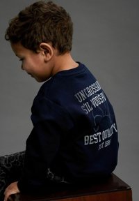 Sweatshirt bleu marine avec un col rond, arborant un texte imprimé en blanc dans le dos. Porté avec un pantalon à motifs sombre, le modèle est assis de côté.