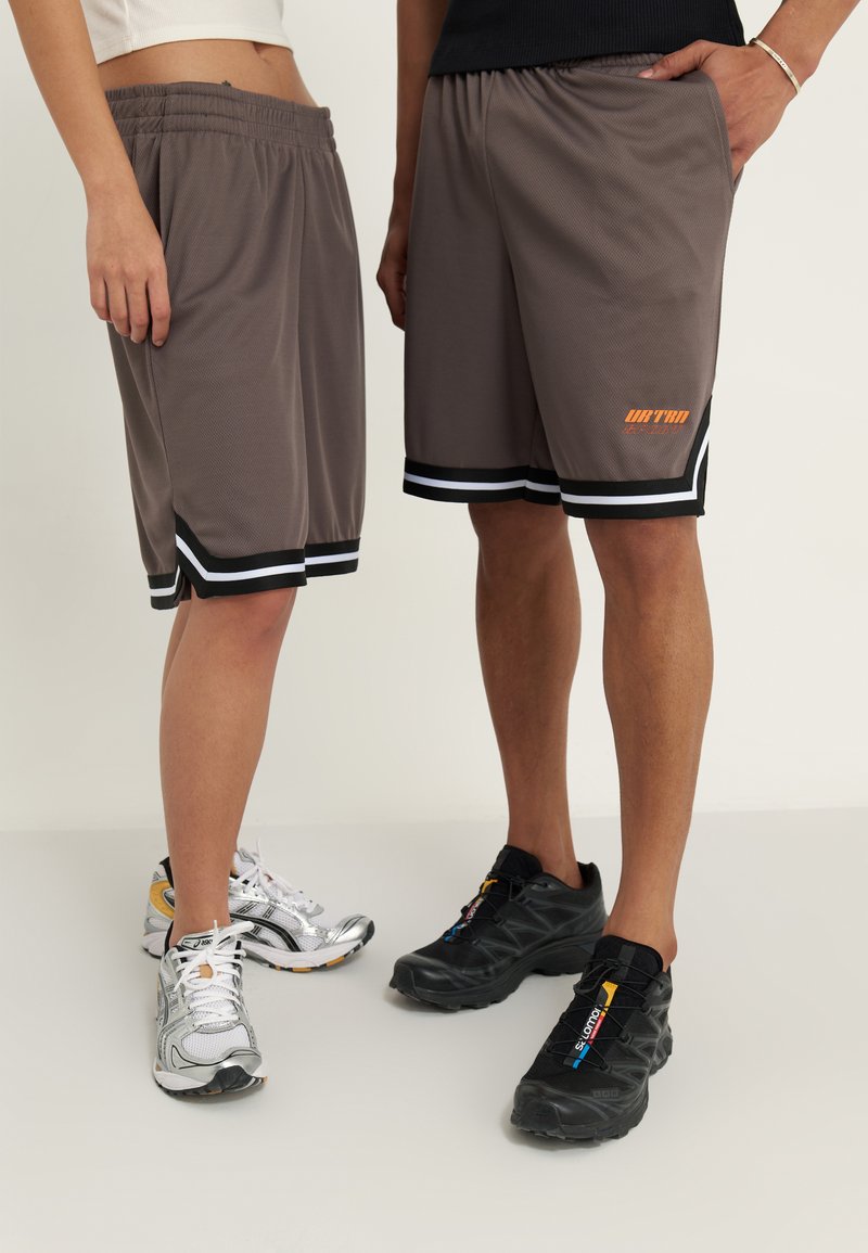Shorts de sport gris avec une bande noire et blanche au niveau de l'ourlet. Présente un logo orange sur un côté. Portés avec des baskets argentées et noires.