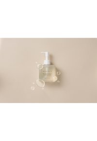 Bottiglia a pompa in vetro trasparente di olio detergente con pompa bianca. L'etichetta presenta "NEEDLY" in un font minimalista. Circondata da morbide bolle, su uno sfondo beige.