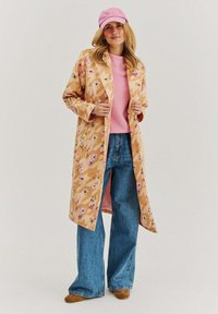 Béžový trenčkot s květinovým vzorem, dlouhými rukávy, světle růžový svetr pod ním, modré široké džíny a růžová kšiltovka.