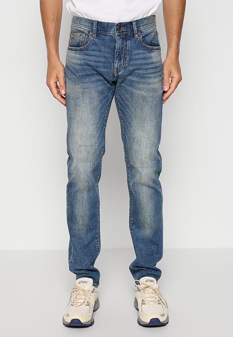 Uomo che indossa jeans blu sbiaditi e sneakers beige ASICS, in piedi di fronte a uno sfondo grigio chiaro uniforme.