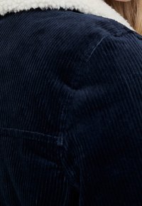 Mörkblå kordjacka med en fluffig vit shearlingskrage. Texturen är ribbad och har synliga sömmar samt en lätt glans.