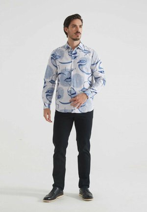 Chemise à manches longues blanche avec des motifs abstraits bleus, dotée d'un col boutonné et de fermetures à boutons. Associée à un pantalon foncé et des chaussures noires.