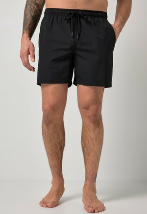 Mężczyzna noszący czarne kąpielówki ze sznurkiem, stojący boso, z widocznym tatuażem na przedramieniu, na jasnym, jednolitym tle.