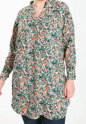 Vrouw draagt een tuniek met lange mouwen met een kleurrijk paisleypatroon in groene, oranje, roze en beige tinten, gecombineerd met een donkere broek.