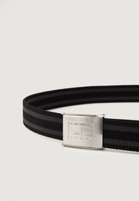 Čierny tkaný opasok s šedými pruhmi a striebornou obdĺžnikovou prackou s gravírovaním "TOMMY JEANS." Textúra je hladká a odolná.
