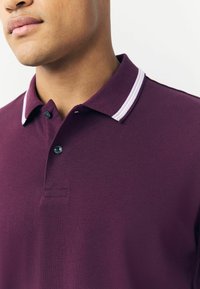 Polo shirt di un viola scuro con un tessuto strutturato, caratterizzato da un bottone a due e un colletto con dettaglio a strisce bianche.