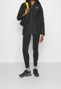 Sort vandtæt jakke med lynlås og hætte, kombineret med sorte leggings og sorte sportssko. Gul rygsæk og grå sokker er synlige.