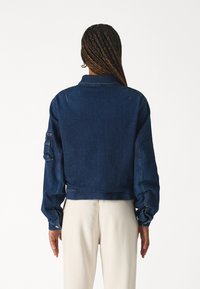 La giacca in denim presenta un colore blu scuro, un design cropped e maniche lunghe con dettagli ai polsini. Il retro non mostra pattern o componenti visibili.