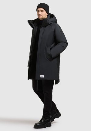 Černá péřová bunda s kapucí, předním zipem a bočními kapsami. Nositá přes černý outfit, který má štítek na spodním lemu a hladkou texturu.