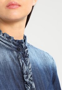 Denimskjorta med volangkrage och knapplist, i en medelblå tvätt. Har en knäppning fram och en mjuk textur.