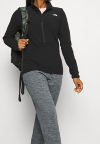 Svart zip-up fleecejacka med hög krage, lättviktsmaterial, bärs tillsammans med grå mönstrade sweatpants och en kamouflagemönstrad ryggsäck.