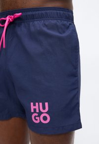 Σκούρα μπλε μαγιό βερμούδα με ροζ κορδόνι και ροζ λογότυπο "HUGO" στο αριστερό μηρό, φορεμένο από άτομο με σκούρο δέρμα.