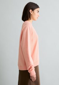 Junge Frau mit kurzen dunklen Haaren, die einen hellrosa Pullover und einen braunen Rock trägt, seitlich stehend vor einem schlichten hellgrauen Hintergrund.