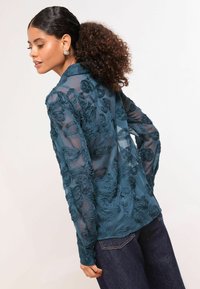 Priesvitná tyrkysová blúzka s kvetinovou výšivkou, dlhými rukávmi a voľným strihom, doplnená o tmavé džínsy. Vyznačuje sa podrobnou textúrou a zložitými vzormi.