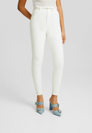 Woman wearing high-waisted white skinny jeans paired with light blue pointed-toe block heels with double buckles.