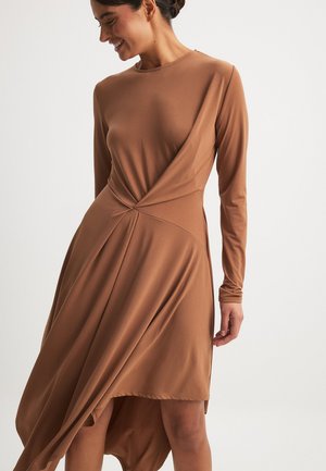 Langærmet brun kjole lavet af glat stof, med et snoet design ved taljen og en asymmetrisk kant med flydende draperinger.