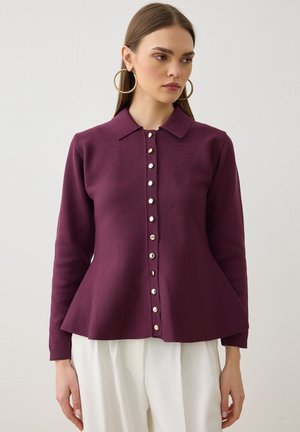 Mujer con una blusa morada ajustada de manga larga con botones y cuello, y pantalones blancos, de pie contra un fondo claro y liso.