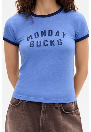 Kobieta w niebieskiej dopasowanej koszulce z ciemną lamówką i napisem "Monday Sucks" z przodu, w połączeniu z brązowymi spodniami.