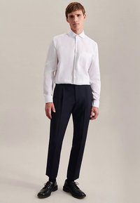 Camicia bianca aderente a maniche lunghe con colletto e taschino frontale, abbinata a pantaloni neri su misura e scarpe nere con lacci.