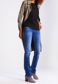 Olijfgroen trenchcoat over een zwarte top, gecombineerd met blauwe jeans en lichtgrijze blokhaklaarzen. De outfit heeft een relaxte silhoutte.