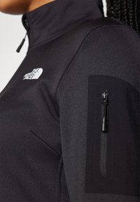 Black zip-up jacket with a high collar, featuring a small white logo on the chest and a zippered pocket on the left arm. Smooth texture.