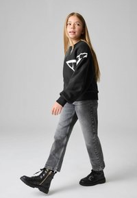 Svart sweatshirt med vit logodetalj, tillsammans med grå jeans och svarta stövlar med guldaccenter och dragkedjor. Släta tygtexturer.