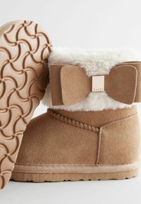 Beżowe zamszowe botki za kostkę z puszystym białym wykończeniem, dużą kokardą z tyłu oraz gumową podeszwą o teksturze z okrągłymi wzorami.