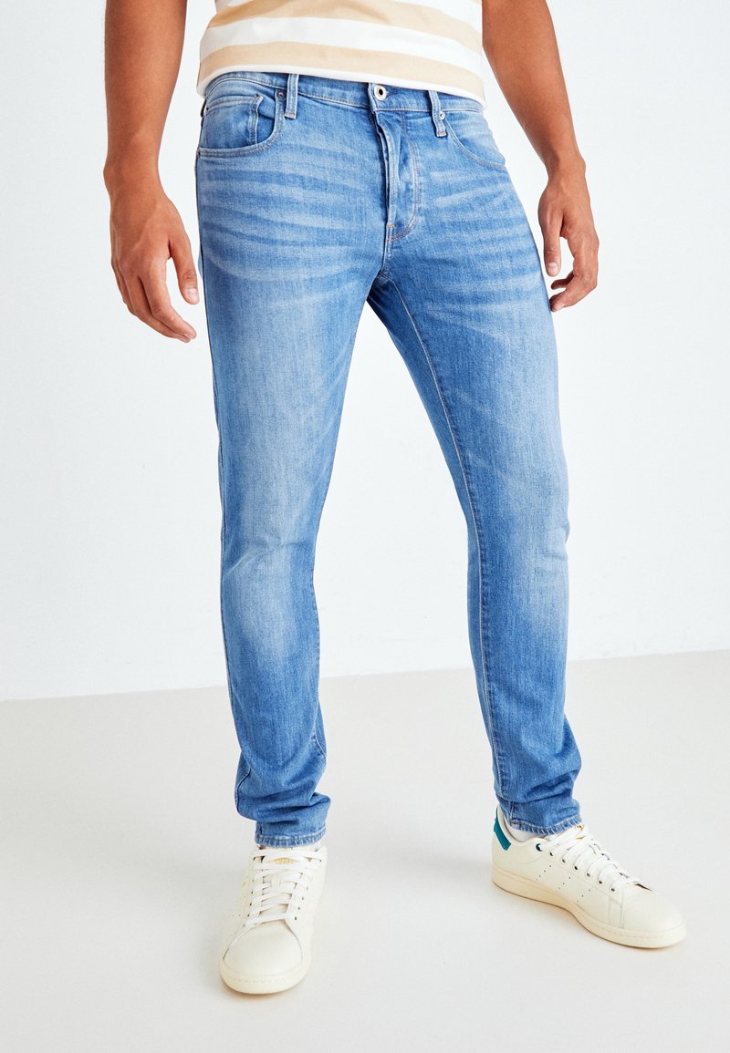 Man som bär ljusblå slim-fit jeans och vita sneakers, stående mot en enkel vit bakgrund.