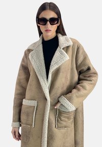 Abrigo de shearling marrón con un cuello y puños de felpa blanca, que presenta dos bolsillos frontales y un diseño de corte relajado.