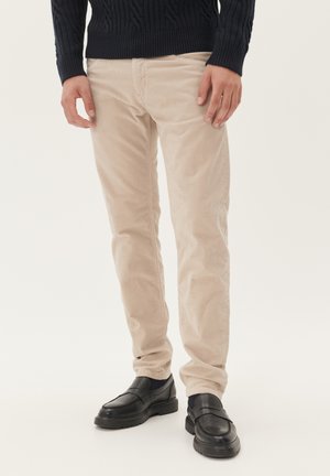 Beige corduroy-housut, joissa on kapea malli, pehmeä rakenne ja hillitty ribbikuvio, yhdistettynä mustiin loafer-kenkiin.