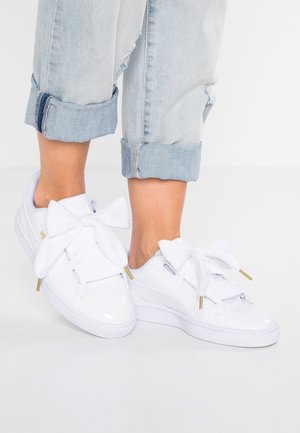 Hvide sneakers med store teksturerede sløjfer på toppen, båret med lyseblå optrukne jeans mod en ensartet hvid baggrund.