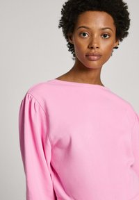 Rozā džemperis ar plašu kakla izgriezumu un pūkainām piedurknēm, izgatavots no mīksta auduma. Tam ir teksturēta virsma un brīvi plūstoša piegriezuma.