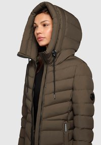 Sieviete valkā brūnu izliekto pūkains jaku ar lielu kapuci un melniem aukliņām virs melna topa, vērsta pa kreisi.