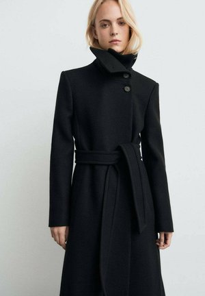 Blondýna v dlouhém černém kabátu s opaskem, vysokým límcem a dvěma knoflíky, stojí před jednobarevným světlým pozadím.