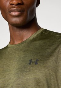 Žalia sportinė marškinė su juodu Under Armour logotipu. Pagaminta iš tekstūruoto audinio su šviesiai tamsiu raštu ir apvalia iškirpte.
