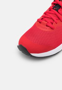 Zapatilla deportiva roja con parte superior de malla, superposiciones texturizadas, suela negra y mediopie blanca. Presenta cordones planos y una puntera acolchada.