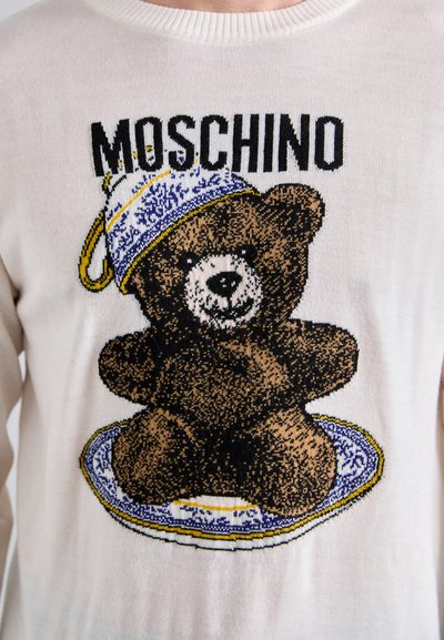 Cremiger Strickpullover mit einem schwarzen Bärenmotiv, der einen blau-weißen Musterteller und eine dekorierte Mütze trägt, beschriftet mit "MOSCHINO."
