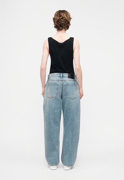 Άτομο με κοντά σγουρά μαλλιά, φοράει μαύρο αμάνικο τοπ και φαρδιά τζιν παντελόνια ανοιχτού μπλε χρώματος, στέκεται με την πλάτη προς τα εμπρός σε απλό φόντο.